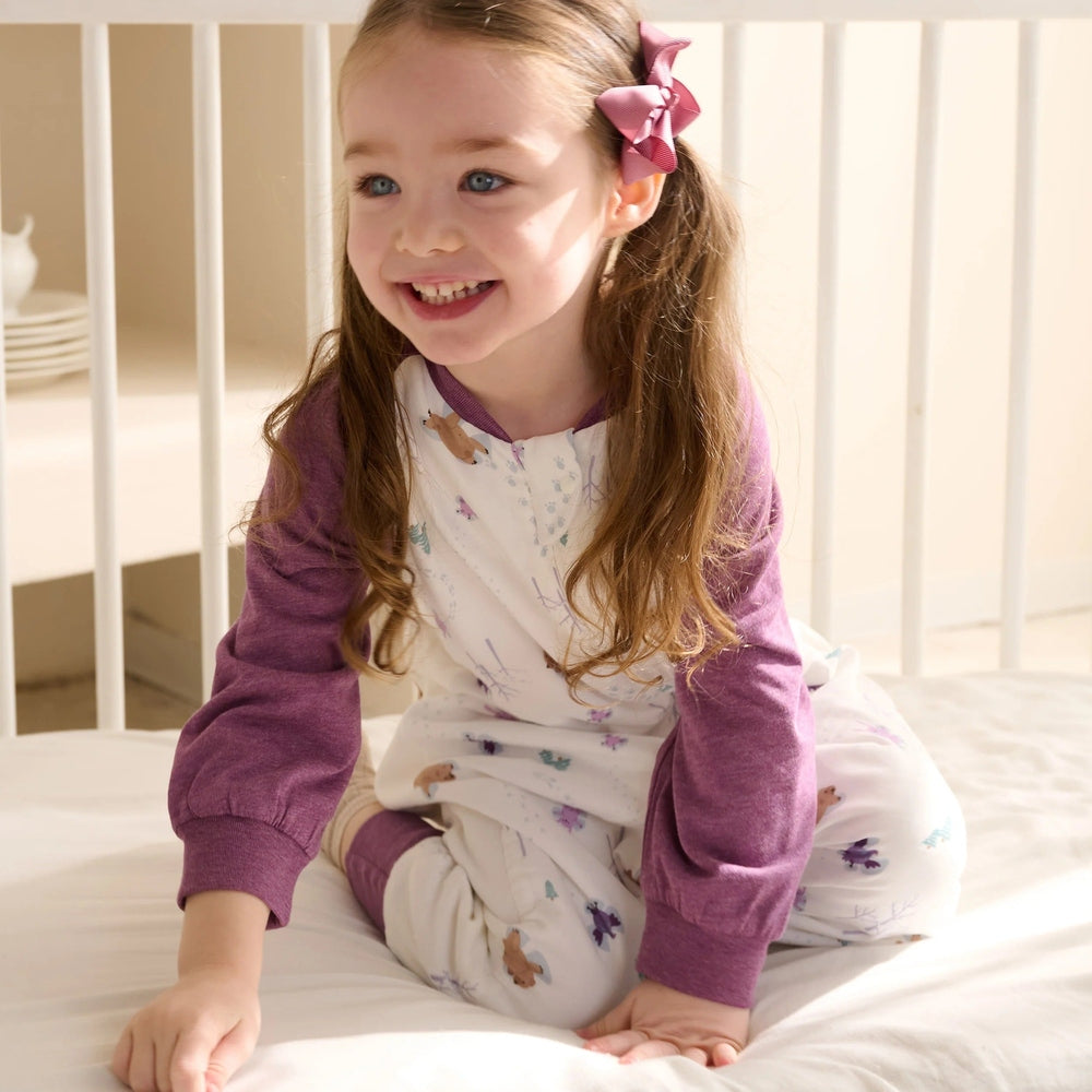 Nest Designs Bamboo Long Sleeve Footed Sleep Bag 1.0 Tog Snow Angels in white with purple sleeves; smiling girl sits on a bed.