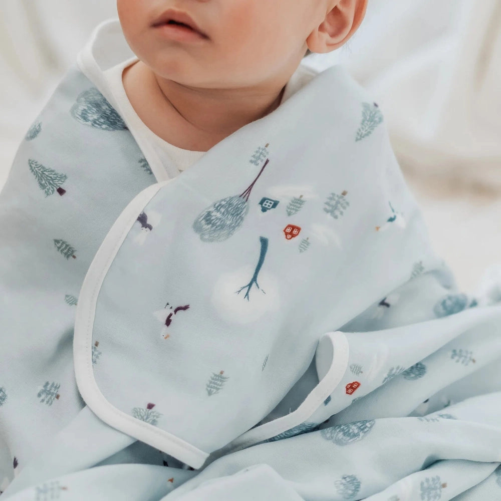 Nest Designs Bamboo Muslin Small Nesting Blanket - baby swaddled in pale blue blanket with white trim and forest pattern.