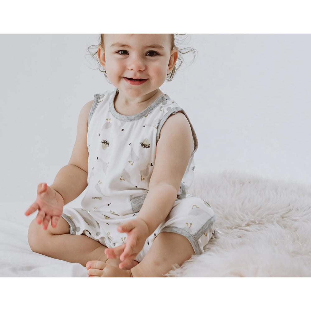 Nest Designs Bamboo Pima Sleeveless Romper The Goose & The Golden Egg: toddler smiles while sitting cross-legged in white romper with gray trim.