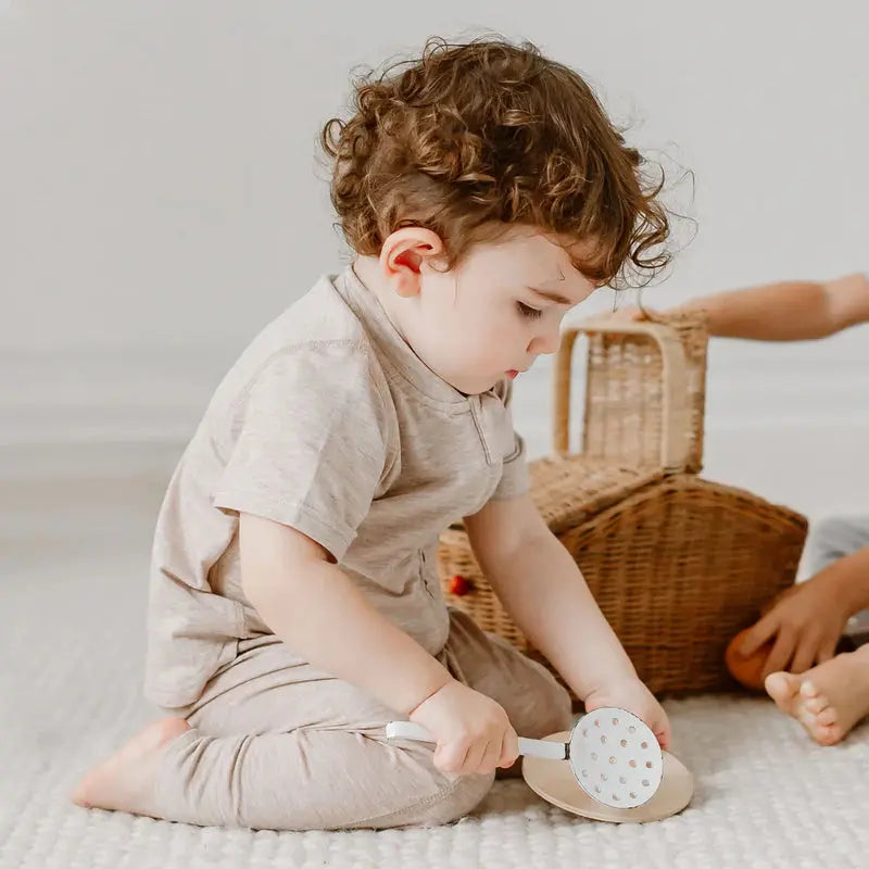 Nest Designs Basics Bamboo Cotton Short Sleeve Drop Bottom Romper in warm taupe; toddler sits on a rug, showcasing soft fabric and easy-access drop bottom.