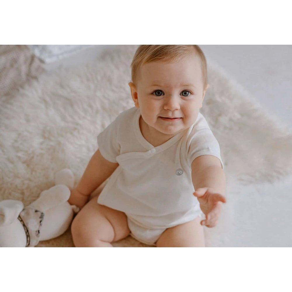 Nest Designs Basics Organic Cotton Ribbed Kimono Short Sleeve Onesie (3 Pack) in white; baby sits, reaching forward to show soft fabric.