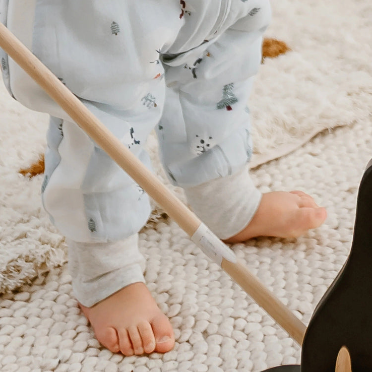 Nest Designs Raglan Bamboo Footed Sleep Bag Snowy Snuggles; baby in light blue footed sleeper with tiny trees, on a plush rug.