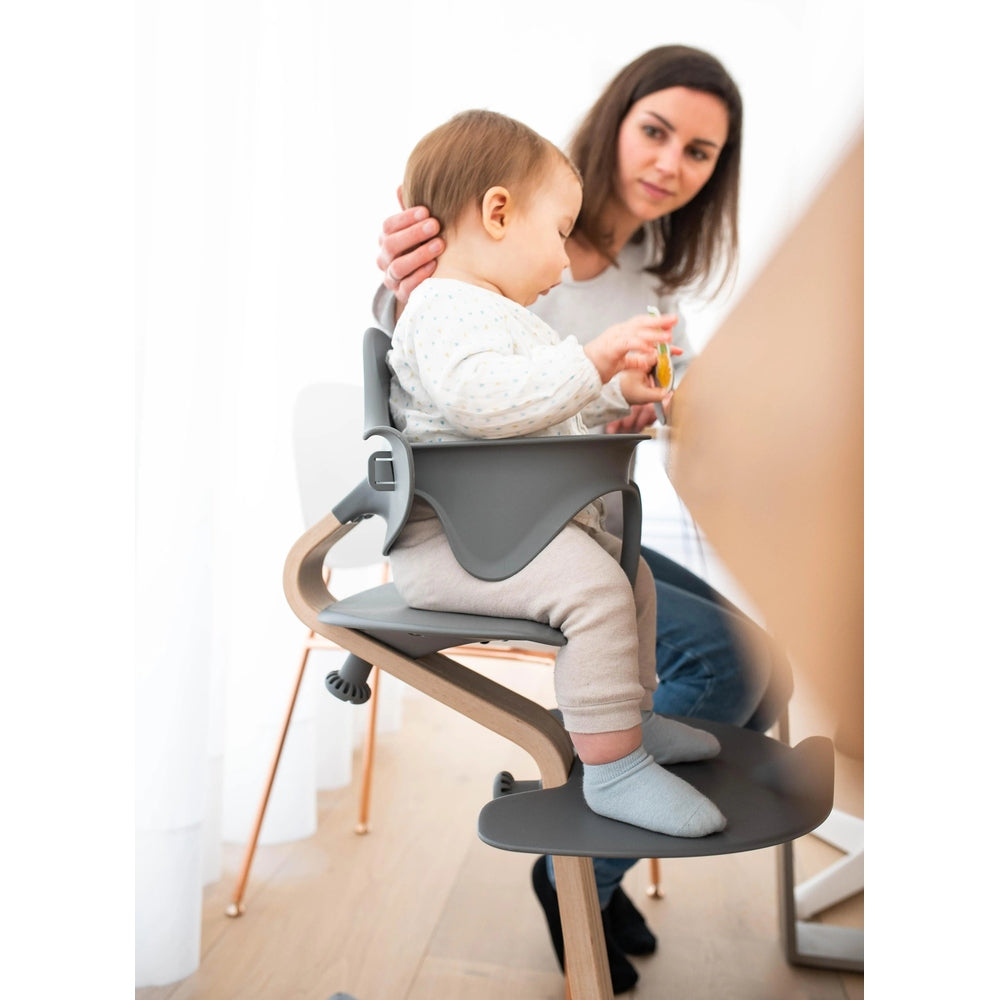 NOMI Highchair White Oak with grey tray; toddler seated as attentive caregiver feeds them.
