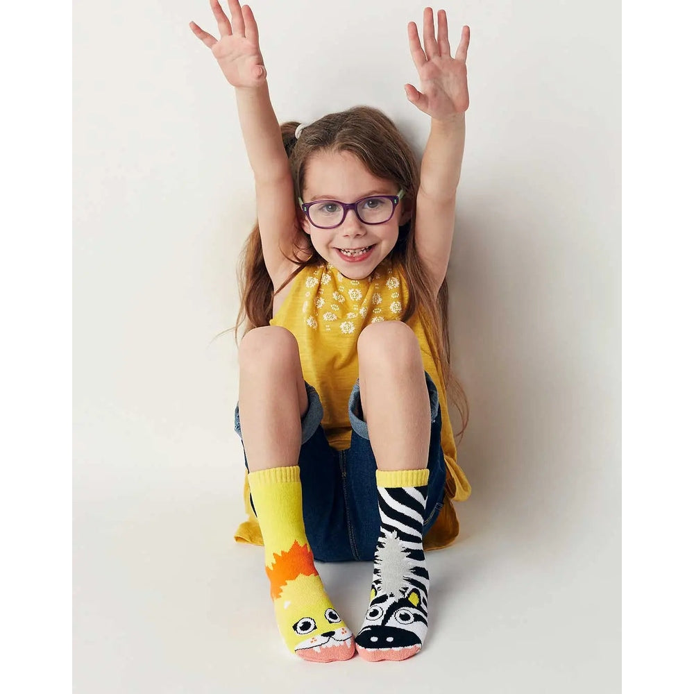 PALS SOCKS Lion & Zebra mismatched socks; yellow lion on left, zebra pattern on right, worn by a cheerful girl raising her arms.