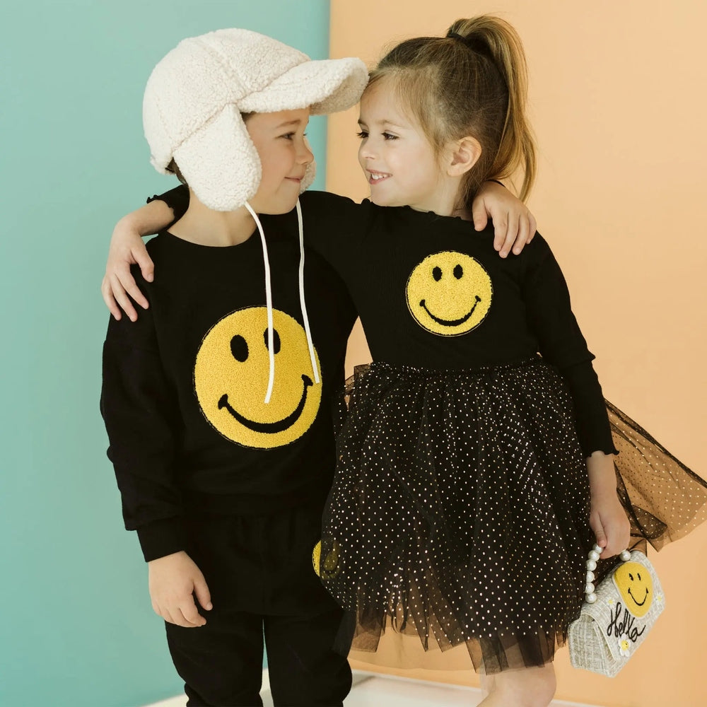 Petite Hailey Smile Tutu Black: two kids hug and smile, one in a black top with a yellow smiley and a dotted tulle skirt.