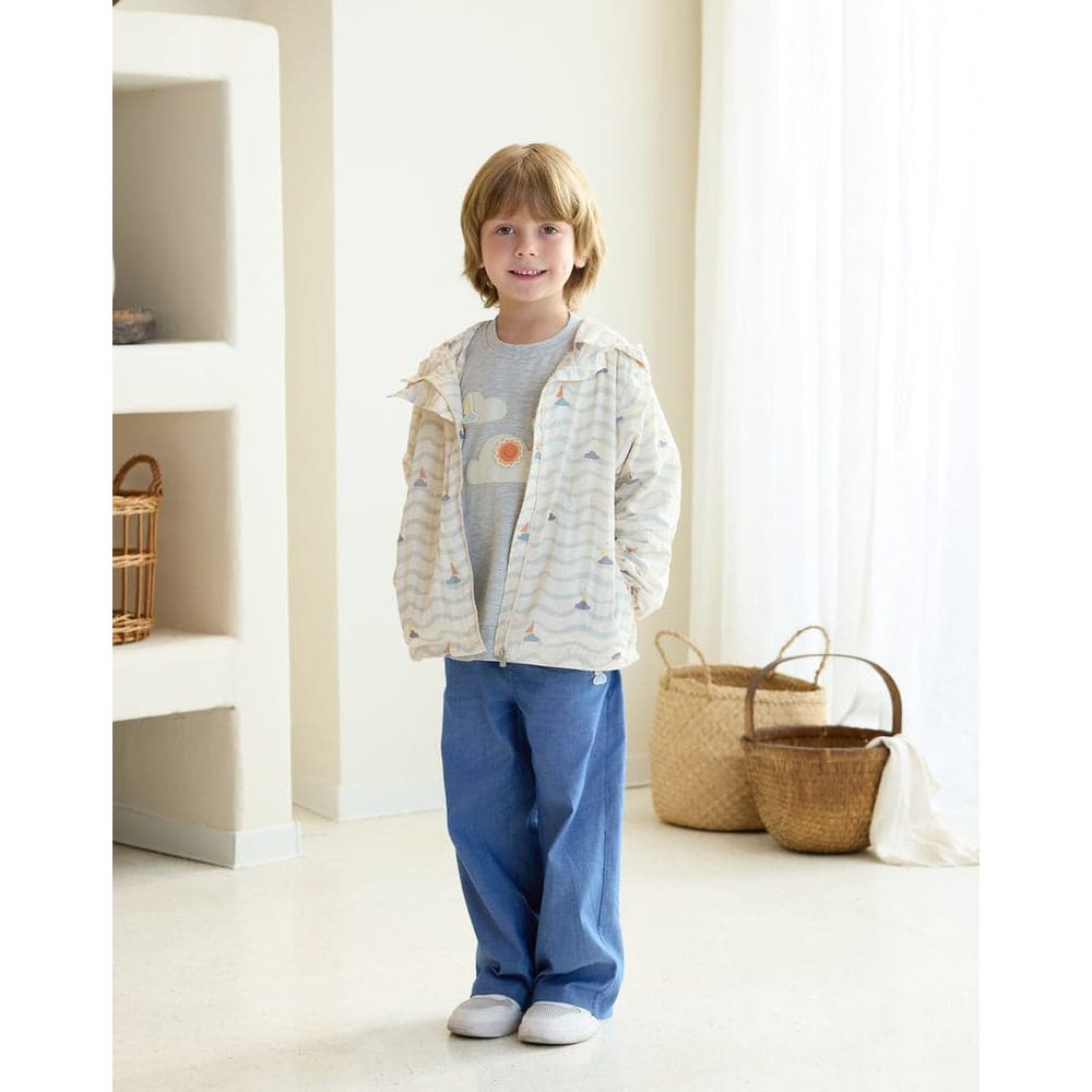 Petite Revery Hooded Windbreaker Sea La Vie worn by a smiling boy in a pale striped jacket, zipper open indoors.
