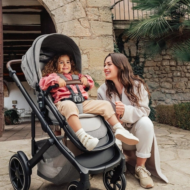 ROMER Tura Stroller in gray with canopy and safety harness; mom chats beside the child seated securely.