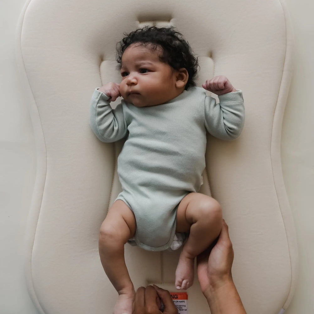 Snuggle Me Organic Lounger Curve baby lounger in beige with a baby in mint-green bodysuit demonstrating the curved snug design.