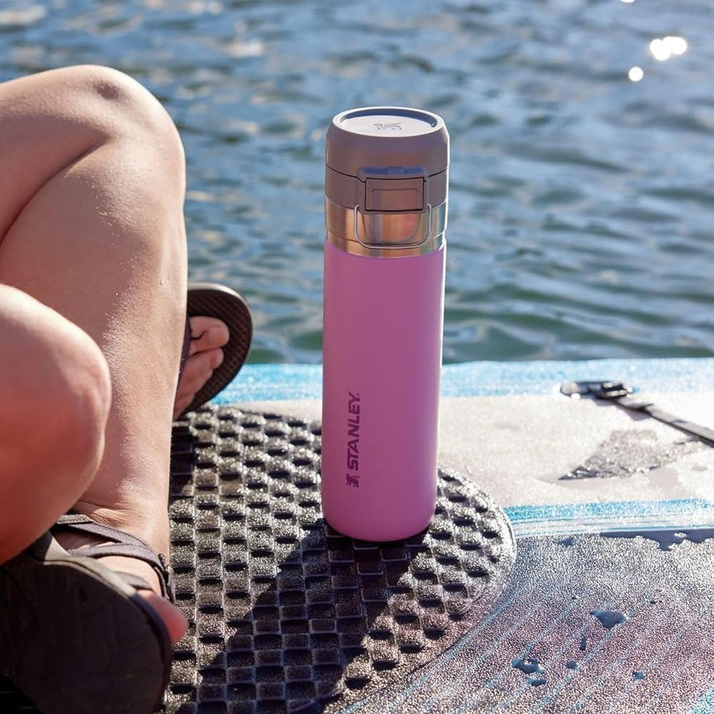Stanley The Quick Flip GO Bottle 24 oz pink with flip-top lid, standing upright on a paddleboard beside a sunlit lake.