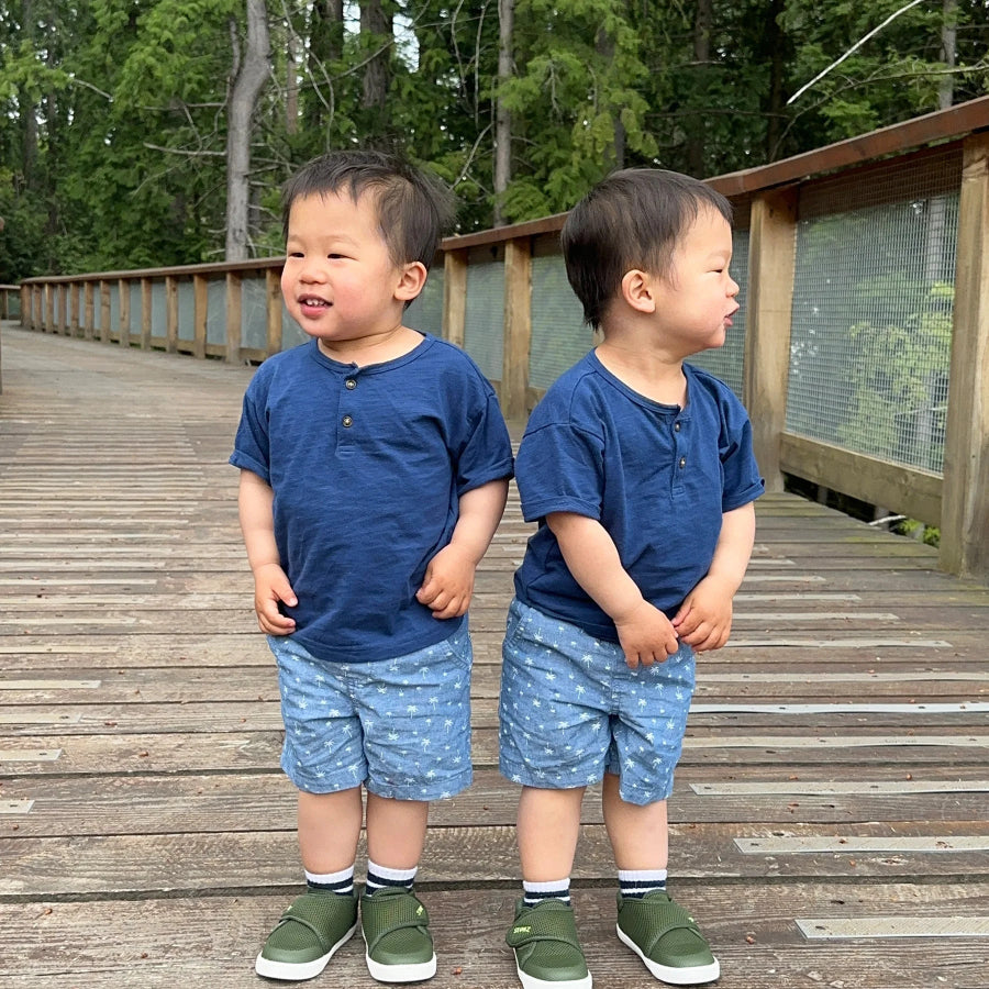 Stonz Cruiser Original Toddler Shoes Cypress olive-green sneakers worn by two toddlers on a wooden boardwalk outdoors.
