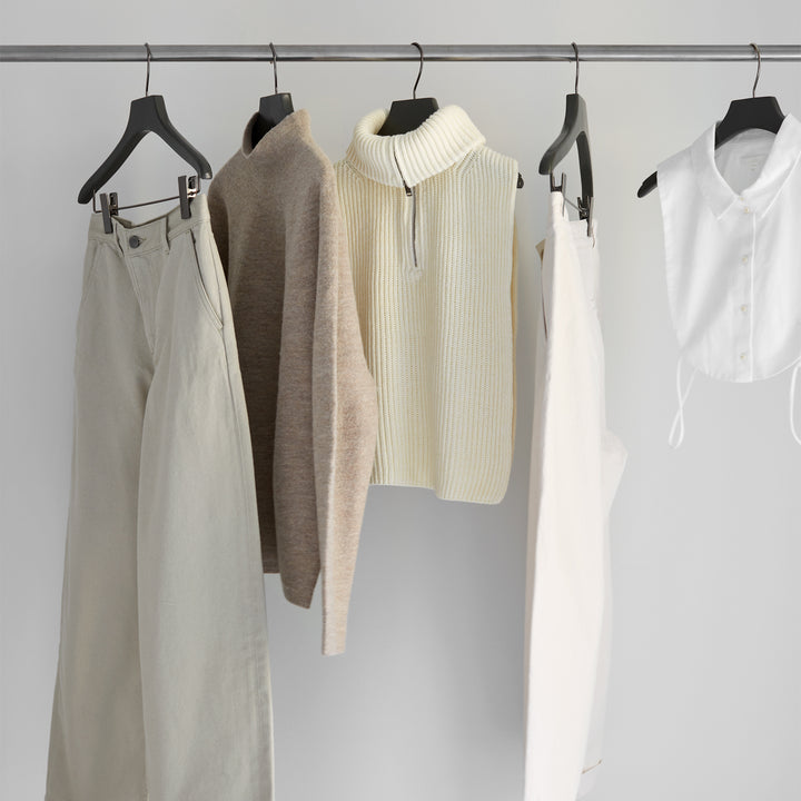 Tangentgc clothing display with five neutral-toned garments on hangers: beige sweater, cream knit, white pants, and white shirt.