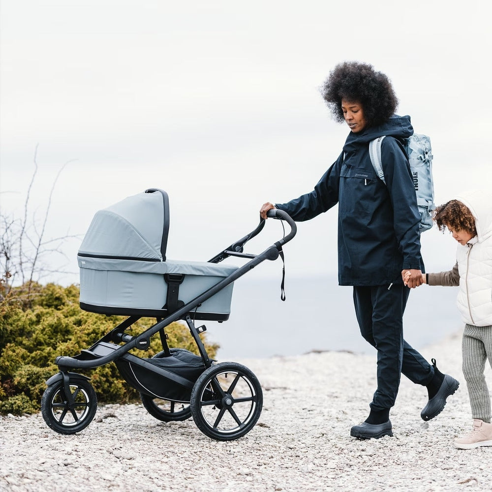 Thule Urban Glide 3 stroller in light gray with large wheels being pushed along a rocky beach by a parent.