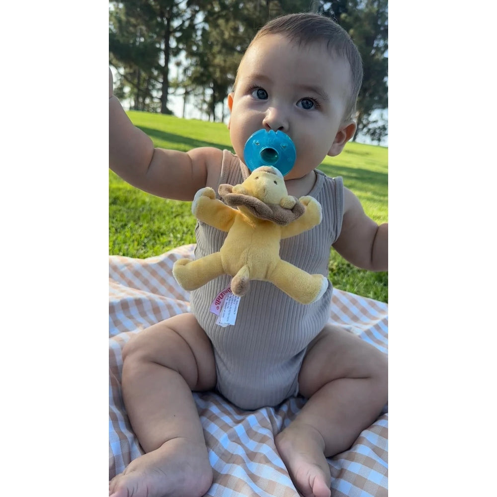WubbaNub Infant Plush Toy Pacifier Lion by Aurora; baby outdoors on a blanket with a blue pacifier attached to a yellow plush lion.