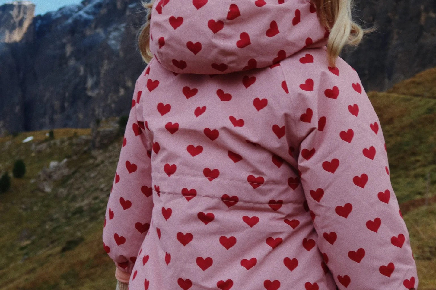 Konges Slojd Mismou Jacket Amour Pink worn by a child walking in a rocky outdoor landscape in a pink heart-patterned puffer with hood.