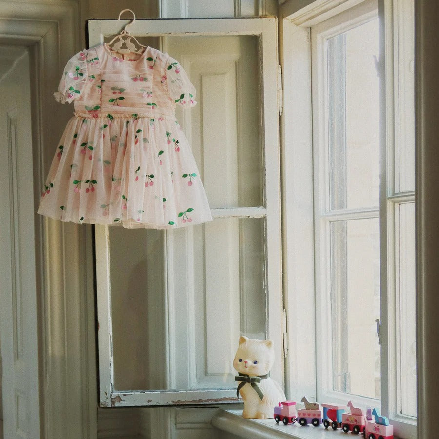 Konges Slojd Tullie Dress Pink Glitter hanging on a hanger by a sunlit window, showing pink tulle skirt.