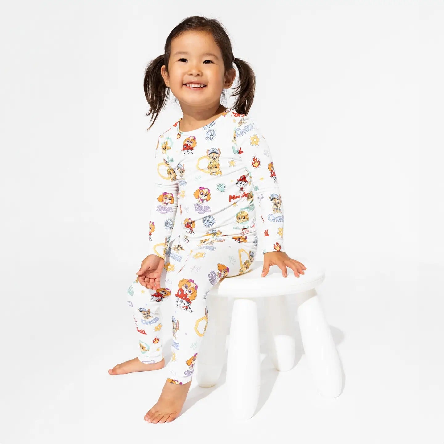 Bellabu Bear Playful Pups Bamboo Kids Pajamas Paw Patrol; smiling girl in white bamboo pajama set sits on a white stool.