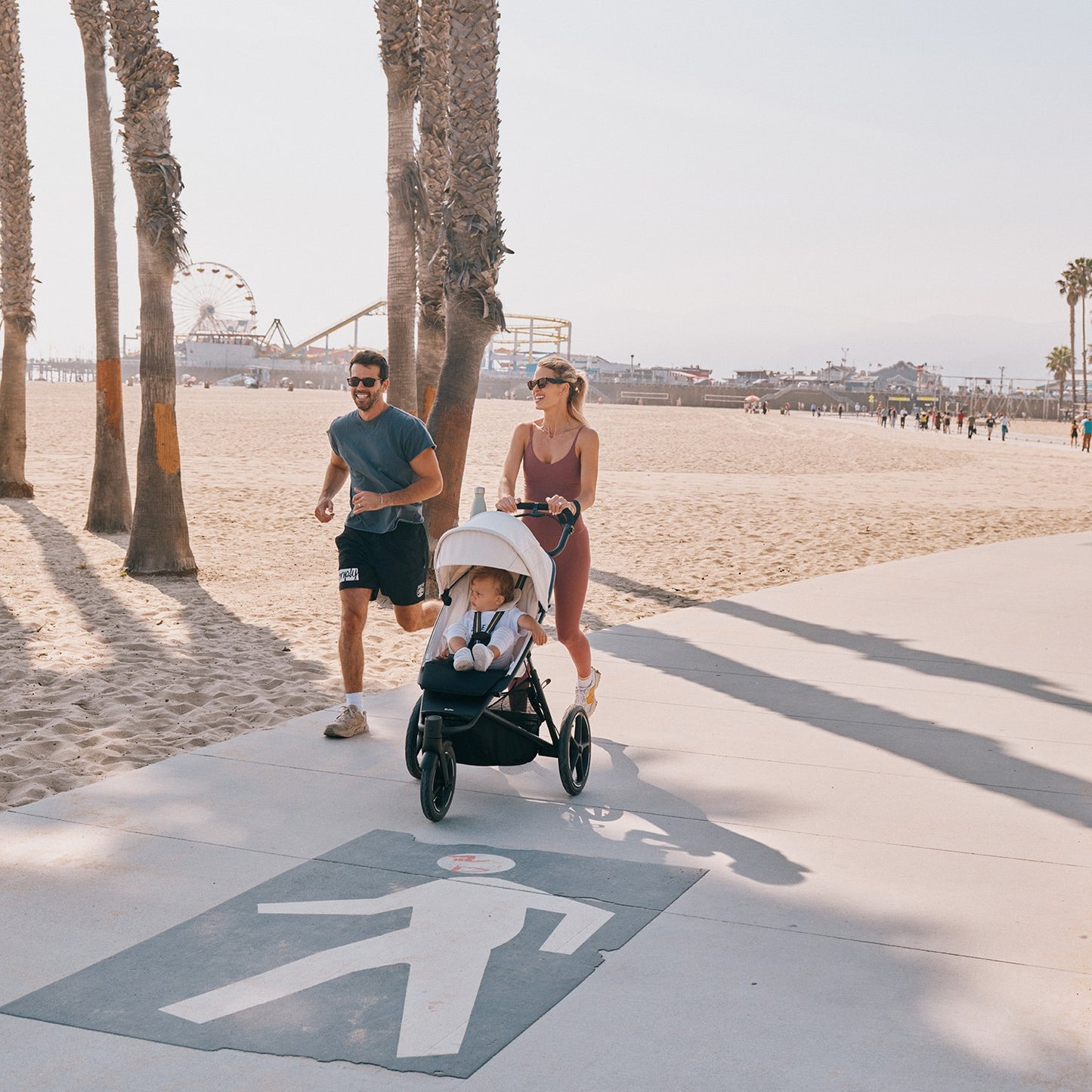 CYBEX Avi Spin All Terrain Stroller on a beach boardwalk, mom pushing with baby as dad jogs nearby.
